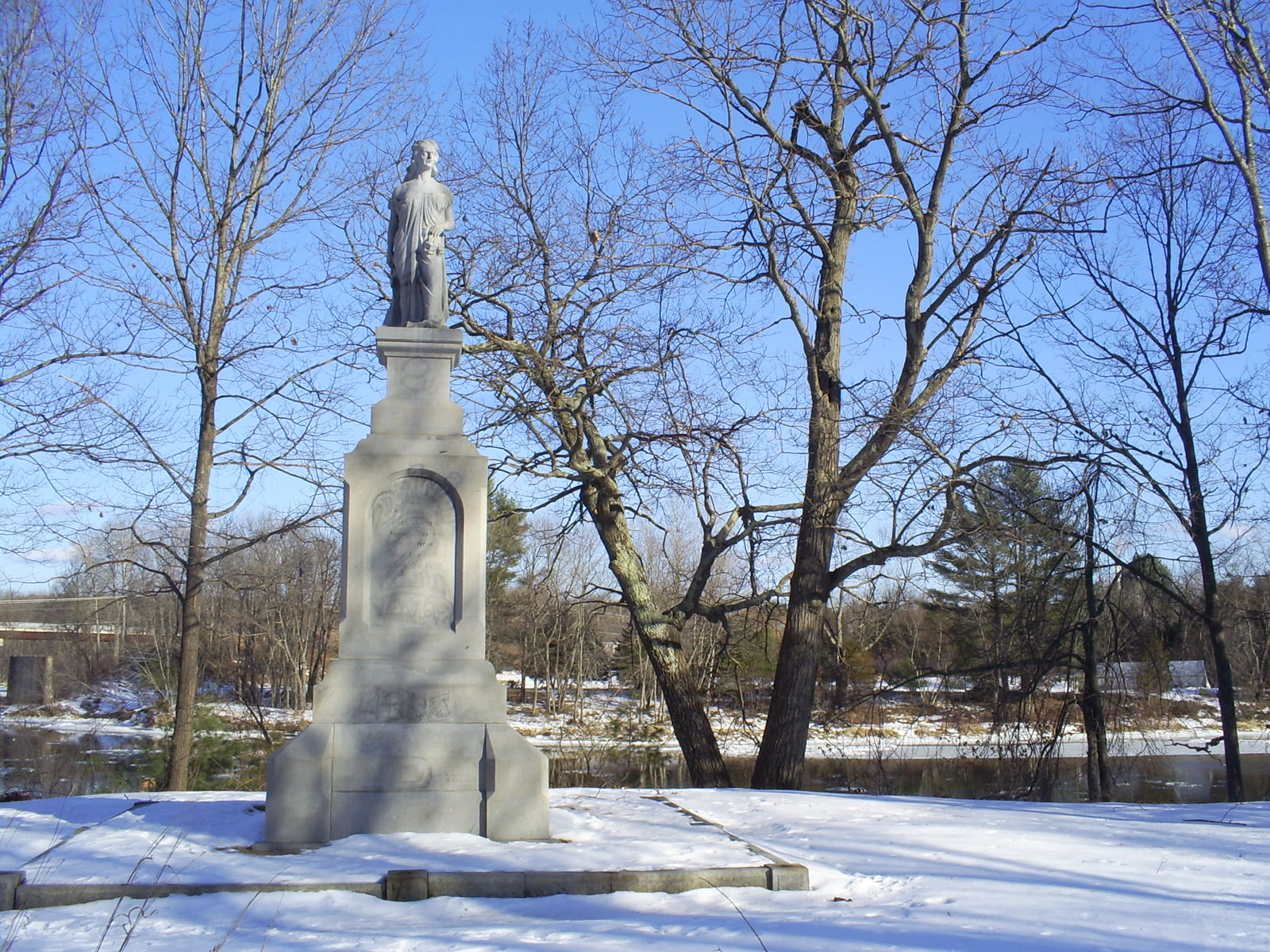 The 1874 Hannah Duston statue at Boscawen, a stone figure on a tall pedestal in snow beside the river, photographed by Craig Michaud.